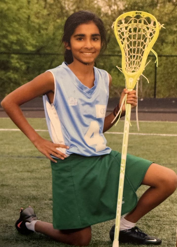 Sejal kneeling in a light blue jersey and green skirt, holding a lacrosse stick